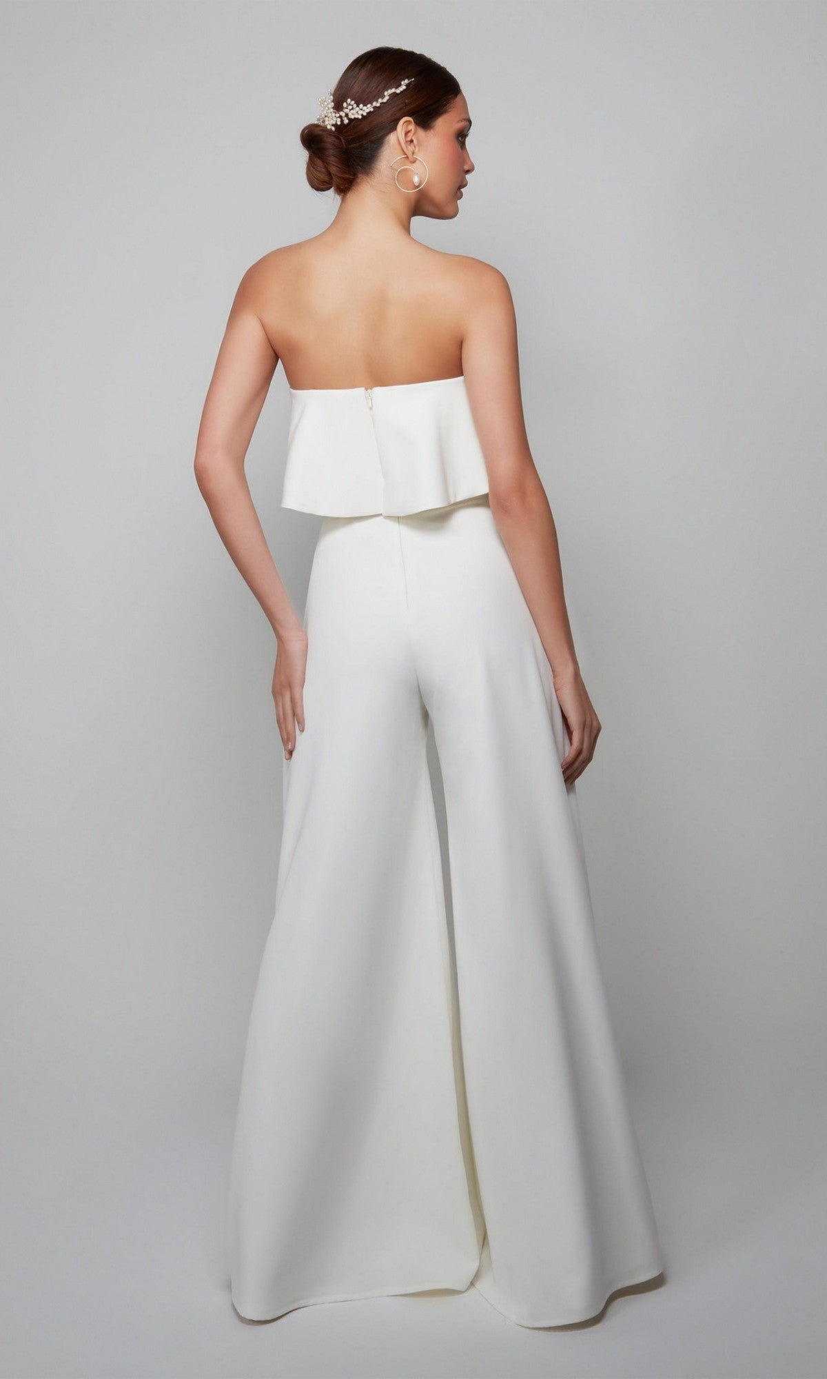 A woman poses with her back to the camera against a plain background, wearing the Long Formal Jumpsuit: Alyce 70015. The strapless jumpsuit features wide-leg pants, a layered top, and she styles her low bun with a decorative hair accessory.
